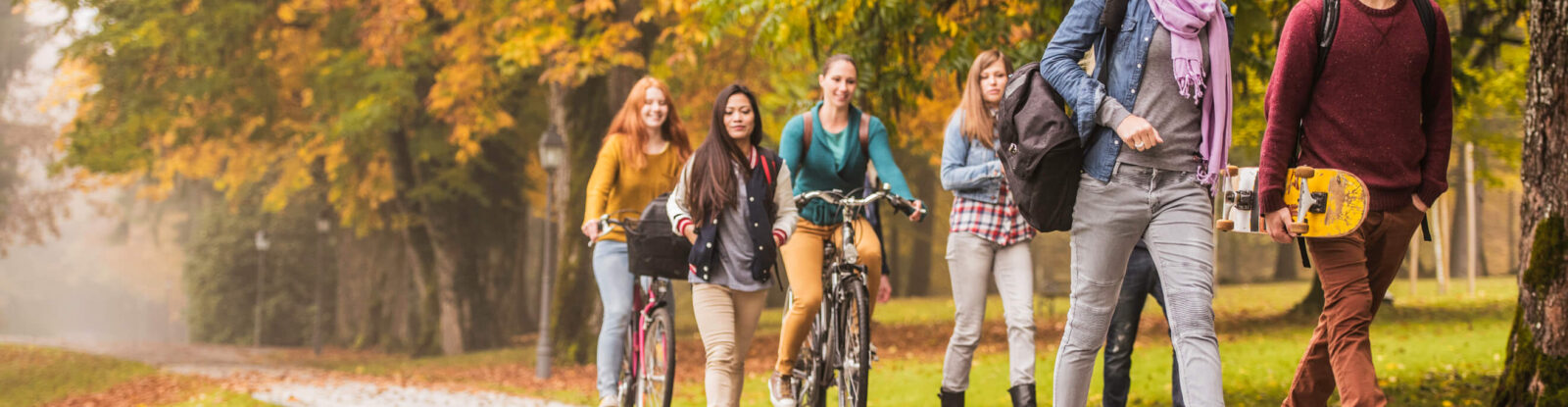 Six college students walking along a path. One is holding a skateboard. Two are riding bicycles. Trees have yellow and orange leaves, with some fallen leaves on the path.