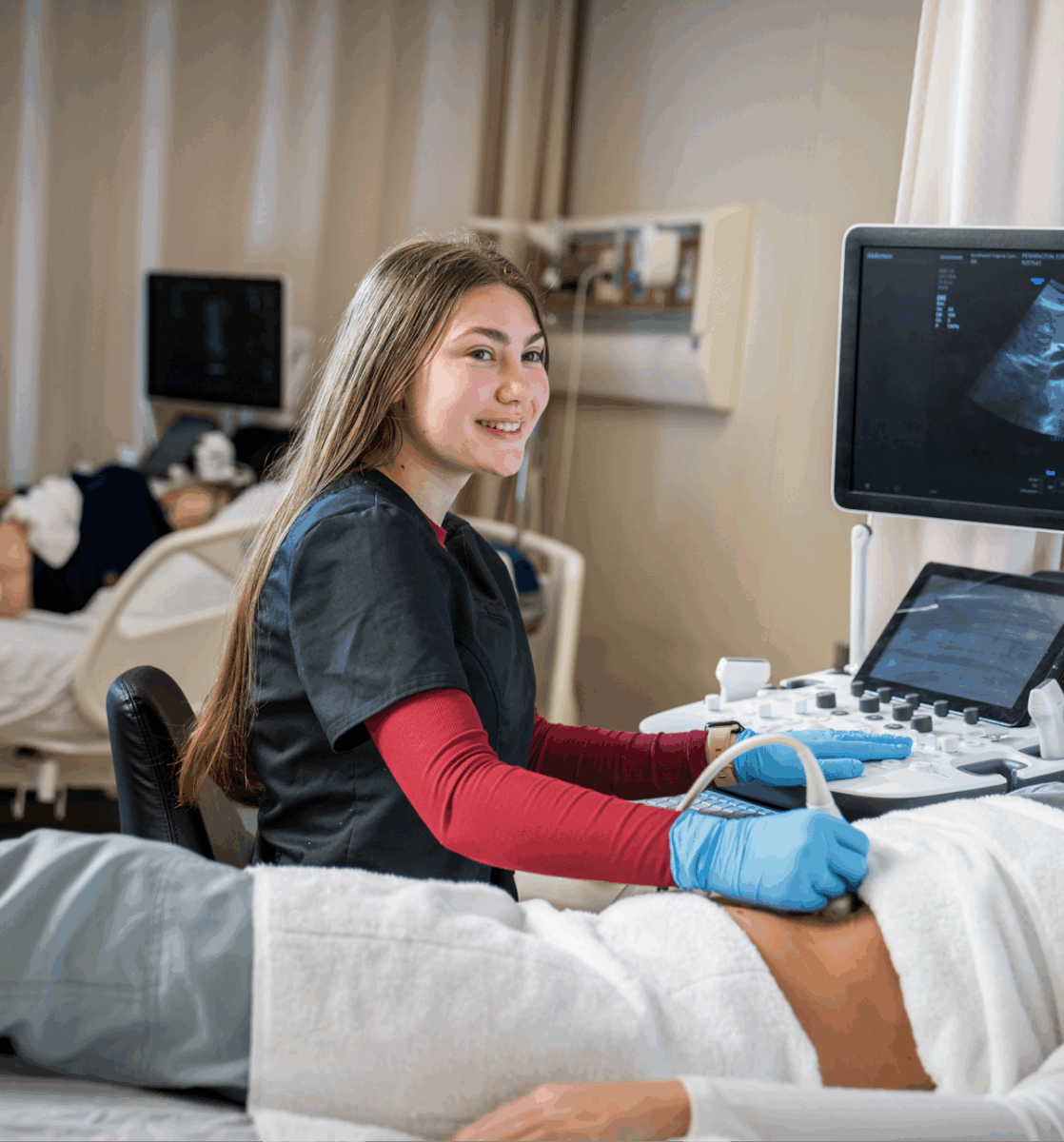 A woman seated next to a patient on a bed holding an ultrasound wand on their stomach.