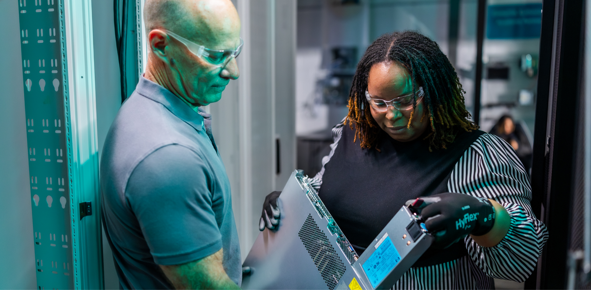 A man and woman standing in a server room. THe woman is pulling a hard drive out of a server the man is holding.