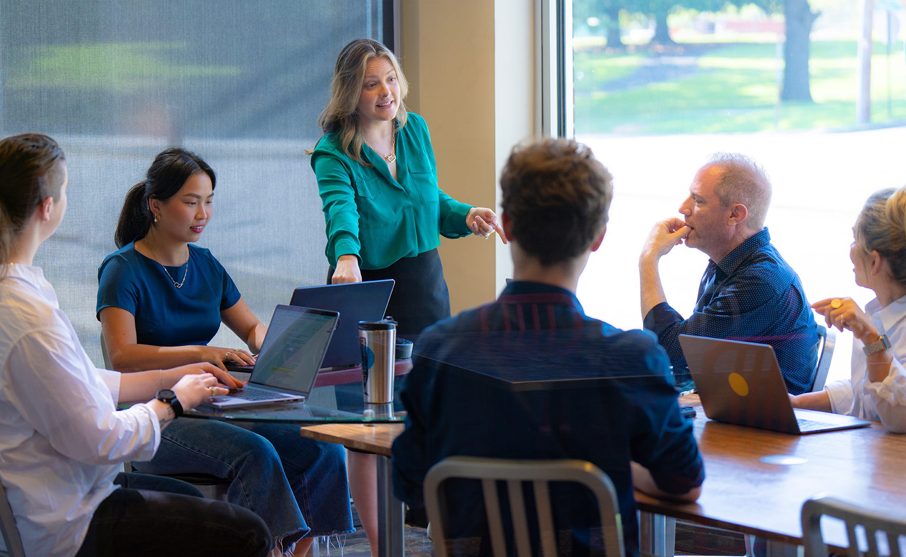A woman standing in a conference room talking to a group of people seated around a table with laptops in front of them.