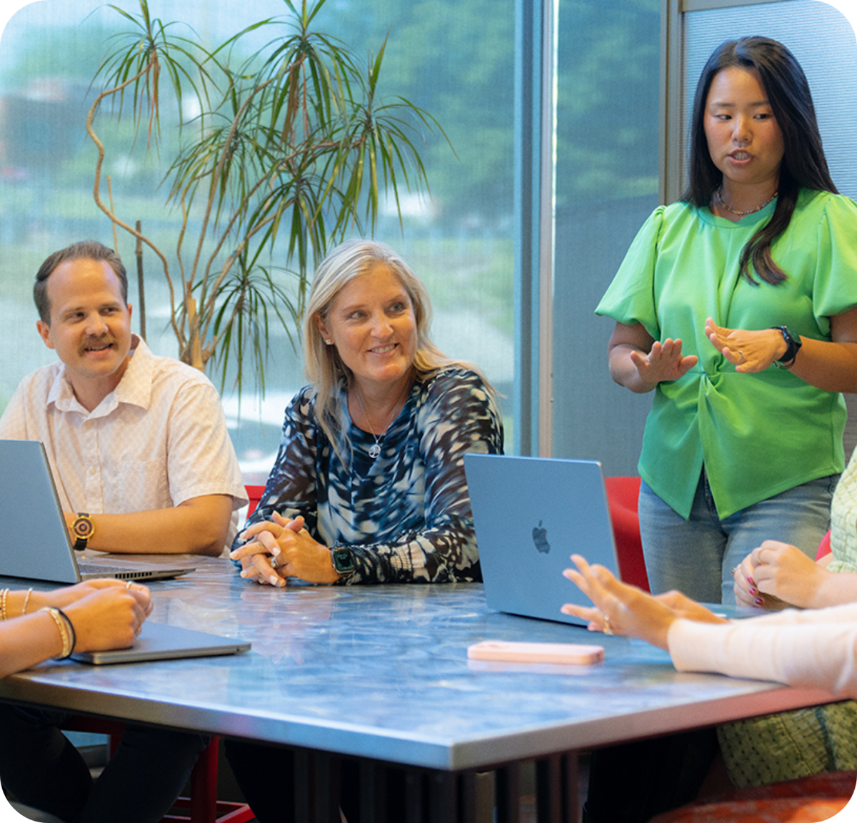 A woman standing in a conference room beside a conference table with others seated around it.