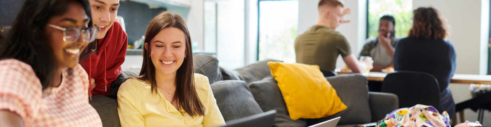 Group of college aged students sitting on a couch looking at a laptop screen. In the background, three additional students are sitting at a table.
