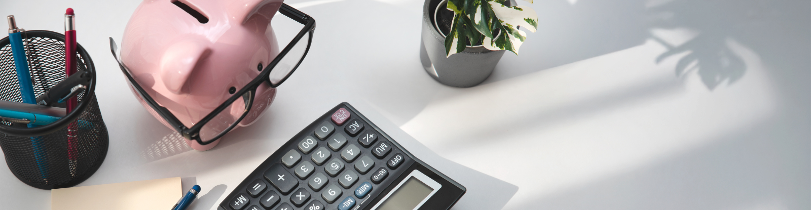 Overhead view of a small piggybank on a desk with a plant, calculator, and pen caddy.